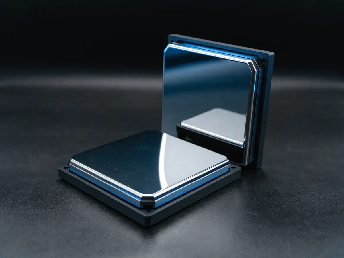 Two oversized square processors in plain black carrier frames, one lying flat and one standing on edge, with blue reflections on their mirrored lids