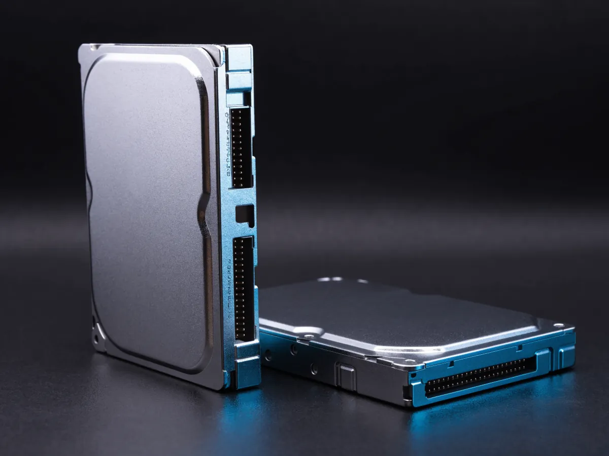 Two 3.5 inch hard disk drives on a dark surface, one standing on its edge showing the connectors and one lying flat behind it with a blank label