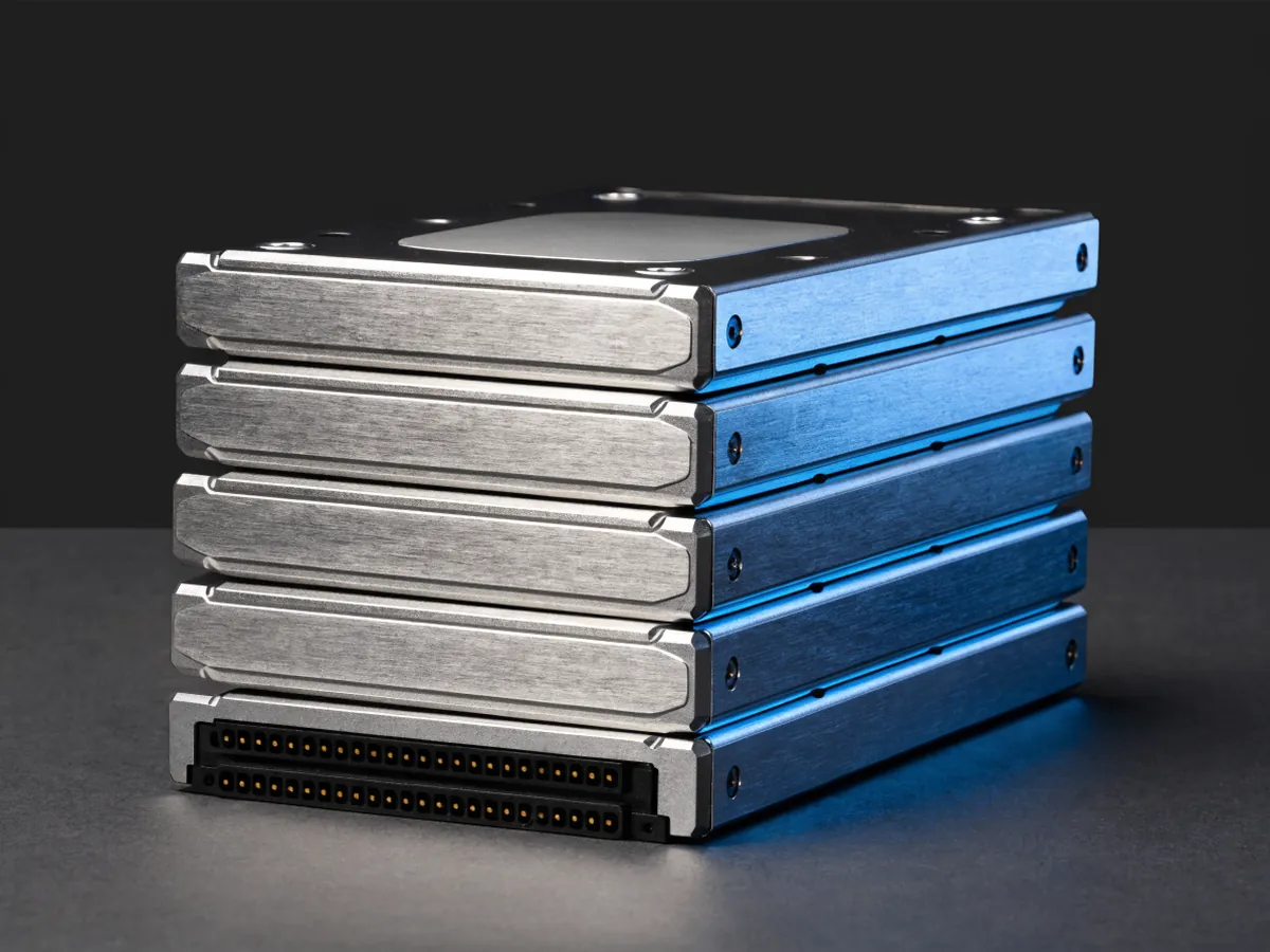 Five 3.5 inch hard disk drives stacked in an offset pile on a dark surface, blank top covers and the connector edge of the front drive visible
