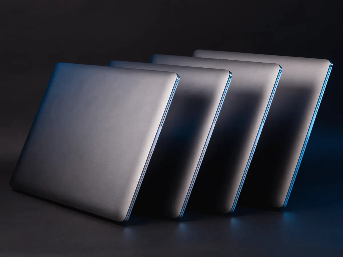 Four identical closed laptops with blank matte lids fanned in a diagonal row on a dark surface under blue rim lighting