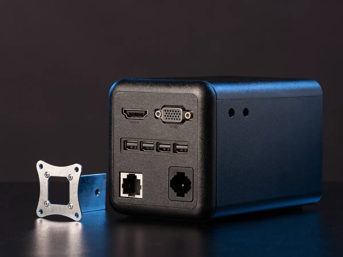 A one litre micro desktop photographed from behind showing its rear port panel, with a flat metal mounting bracket beside it on a dark surface