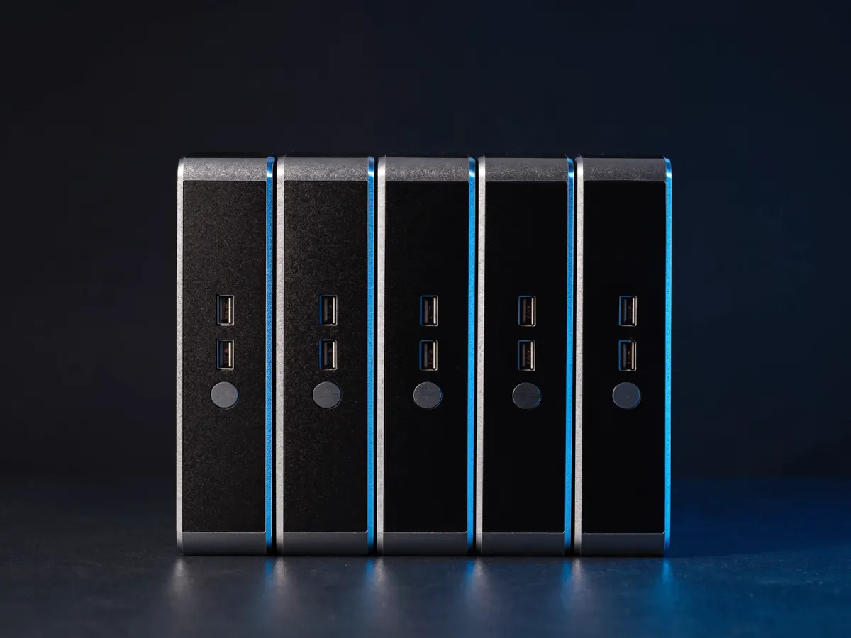 Five identical one litre micro desktop computers standing on edge in a row with front ports facing the camera, lit in cool blue on a dark surface