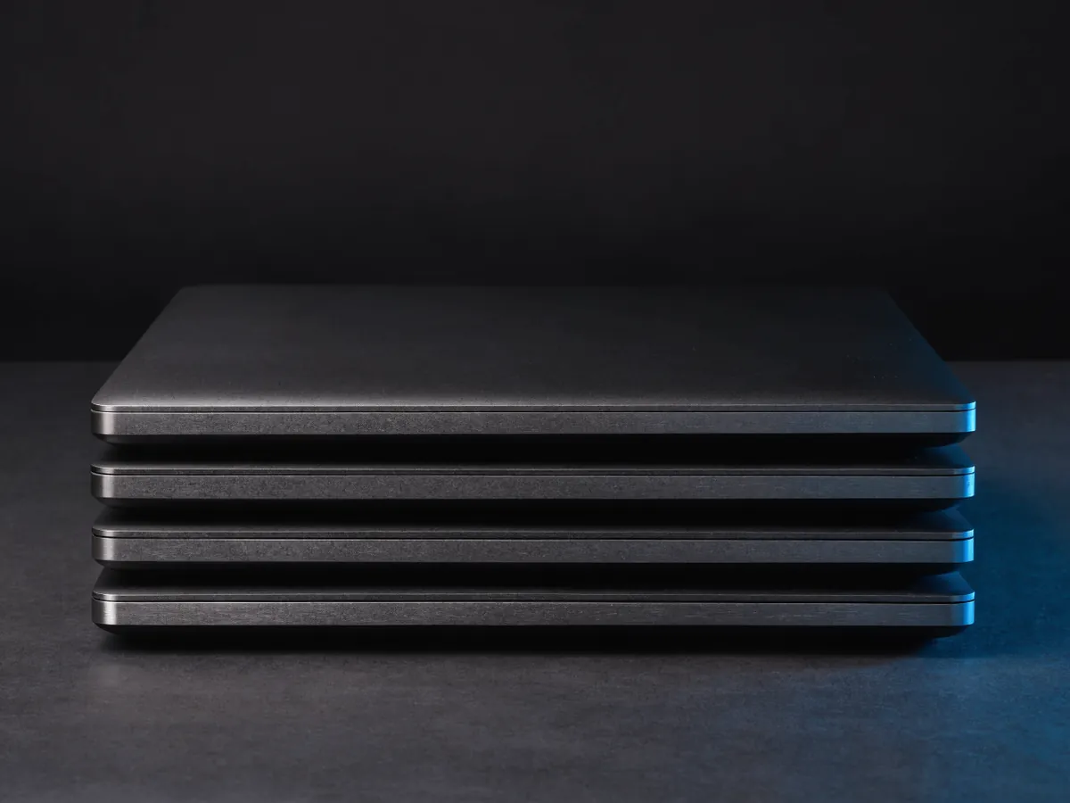 Three matte black laptops stacked flat with lids closed and side ports visible, on a dark surface under cool blue lighting