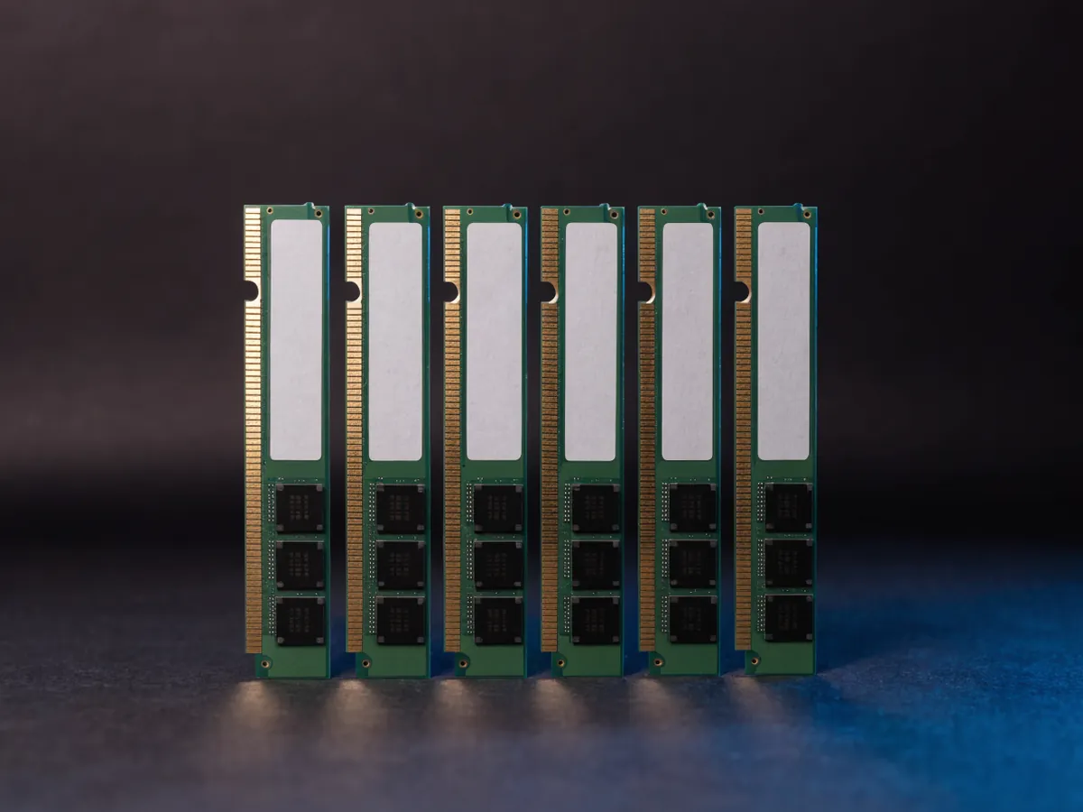 Six full length server memory modules standing upright in a row on a dark surface, showing gold edge contacts and blank unprinted labels under cool blue lighting