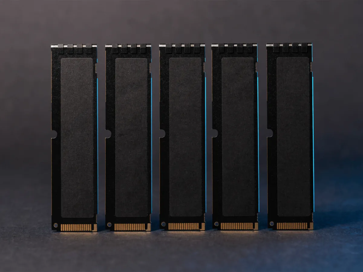 Five memory modules with plain matte black heat spreaders standing upright in an evenly spaced row on a dark surface under blue rim lighting