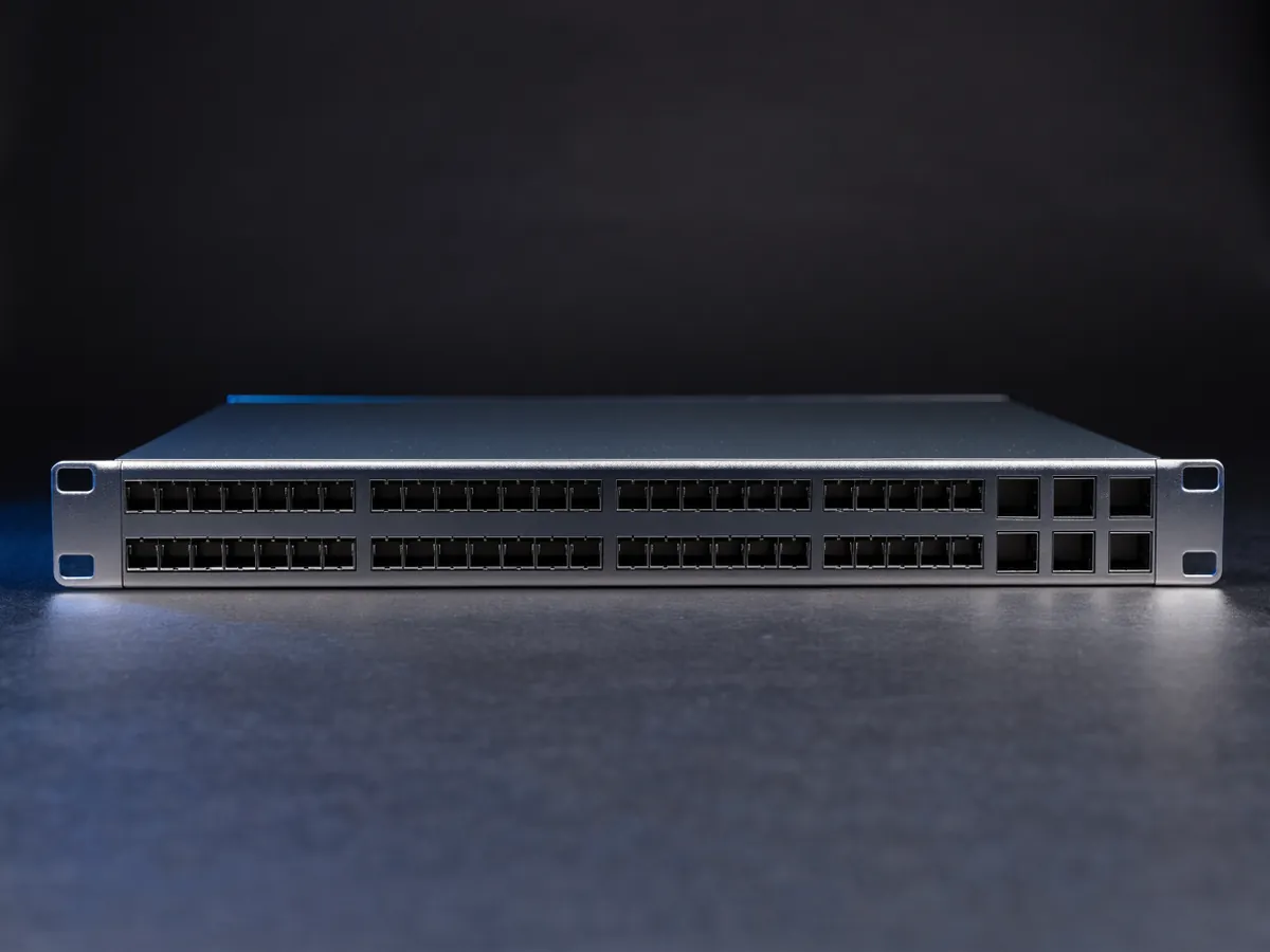 Front elevation of a 1U data center switch on a dark background, showing forty eight small empty transceiver cages in two rows and six larger square cages at one end